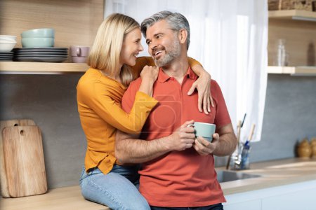 Photo for Pretty blonde woman sitting on kitchen table, hugging her loving husband from behind, happy middle aged couple drinking coffee at home, bonding, having conversation, copy space - Royalty Free Image
