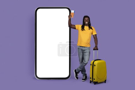 Photo for Happy long-haired young african american man traveller standing next to smatphone with white empty screen, carrying yellow luggage, holding passport with tickets, mockup, purple studio background - Royalty Free Image