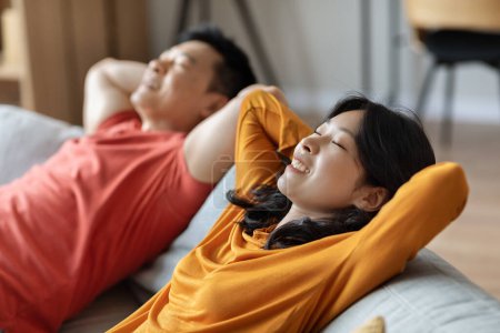 Foto de Relajada pareja asiática relajándose juntos en casa, feliz hombre chino de mediana edad y hermosa mujer joven reclinado en el sofá con los ojos cerrados y las manos detrás de sus cabezas, vista lateral - Imagen libre de derechos