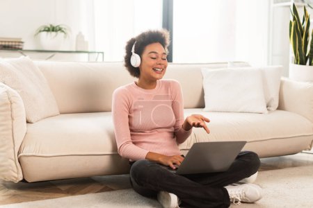 Photo for Cheerful millennial african american woman in wireless headphones has video call on laptop, gesturing sits on floor in bright living room interior. Work, business, study and meeting remote at home - Royalty Free Image