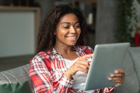 Photo for Glad happy black young woman reading good message, typing on tablet, watching video in living room interior. Offer and ad, new app, social networks, chat and surfing in internet at home with device - Royalty Free Image