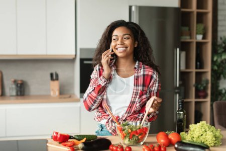 Téléchargez les photos : Préparer maison végétarienne manger. Glad jeune femme noire dans une salade de cuisine décontractée de légumes biologiques, appels par téléphone dans l'intérieur de la cuisine moderne. Communication à distance, commérages et bonnes nouvelles à la maison - en image libre de droit