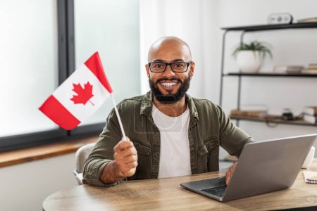 Téléchargez les photos : Heureux latin assis avec le drapeau du Canada et utilisant un ordinateur portable, assis au bureau à la maison. Éducation étrangère moderne en ligne, émigration et citoyenneté, concept de travail à l'étranger - en image libre de droit