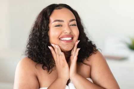 Photo for Portrait of beautiful black chubby woman touching her cheeks and smiling at camera, attractive young lady enjoying skincare treatments at home - Royalty Free Image