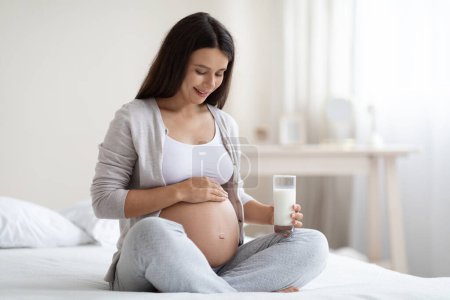 Photo for Happy beautiful young pregnant woman in homewear sitting on bed at home, holding glass of fresh milk and touching her big belly, panorama with copy space. Vitamins and minerals for babys growth - Royalty Free Image