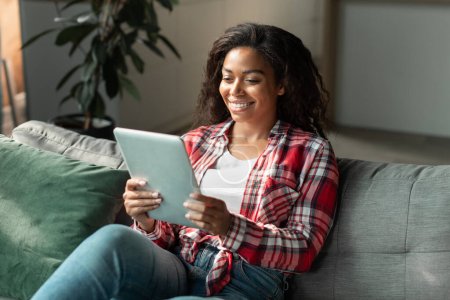 Photo for Cheerful black young woman reading blog, chatting on tablet, watch video, enjoy online shopping in living room interior. New app, social networks for communication, serfing and new normal at home - Royalty Free Image