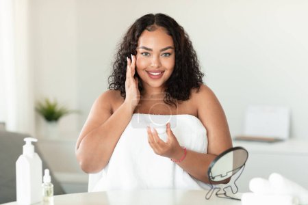 Photo for Portrait of black chubby lady applying face cream, enjoying domestic spa procedure, standing in bath towel at home. Lovely plus size woman taking care of her skin after shower - Royalty Free Image