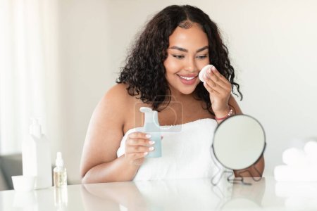 Mujer gordita negro feliz limpiando su cara, utilizando almohadillas de algodón y producto de limpieza, mirando al espejo en el dormitorio y sonriendo. Señora atractiva con tóner facial y almohadilla de algodón, espacio de copia