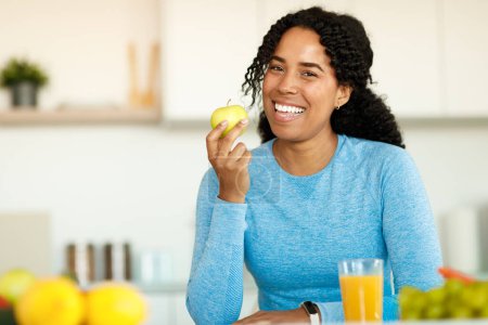 Foto de Retrato de mujer negra excitada en ropa deportiva sosteniendo manzana y sonriendo a la cámara, sentada en el interior de la cocina de luz. Alimentación saludable y nutrición para adelgazar, recetas de alimentos bajos en calorías - Imagen libre de derechos