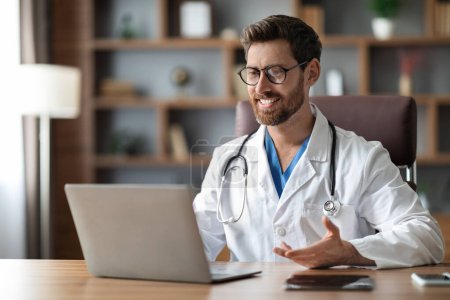 Photo for Online Consultation. Handsome Male Doctor Teleconferencing With Patient Via Laptop, Smiling Physician Man Sitting At Desk And Making Video Call On Computer, Enjoying Telemedicine, Copy Space - Royalty Free Image