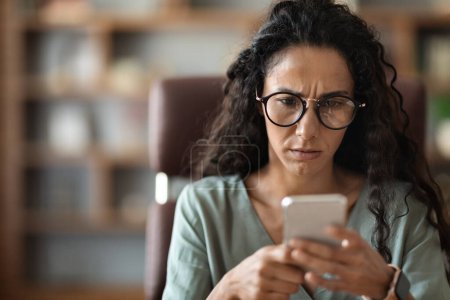 Photo for Closeup photo of anxious woman wearing eyeglasses sitting at workplace, looking at phone screen, reading emails, having difficulties with business, got hurtful message, office interior, copy space - Royalty Free Image