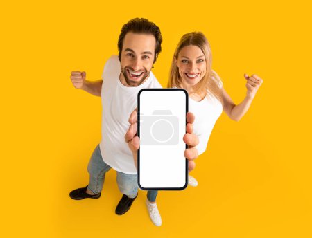Happy millennial caucasian couple in white t-shirts enjoy winning online, show smartphone with blank screen, isolated on yellow background. Huge sale, ad and offer, website, presentation of new app