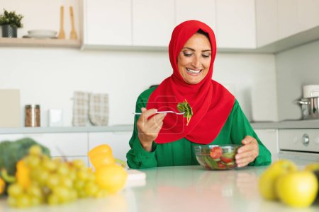 Foto de Alegre señora musulmana madura comiendo ensalada de verduras frescas, sentado en la cocina moderna en casa, espacio para copiar. Nutrición saludable y pérdida de peso concepto de recetas - Imagen libre de derechos
