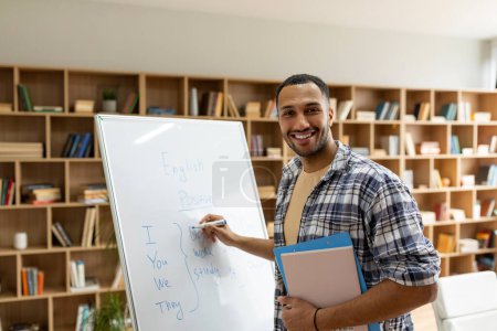 Photo for Happy arab male teacher writing rules on whiteboard, looking and smiling at camera in living room interior. English language lesson, lecture, presenting new topic, education and be safe - Royalty Free Image
