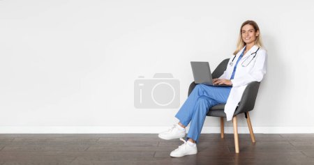 Foto de Banner Médico. Señora sonriente del doctor usando el ordenador portátil mientras que se sienta en la silla contra la pared blanca en el hospital, Médico femenino hermoso que usa uniforme que da consultas en línea vía computadora, espacio de copia - Imagen libre de derechos