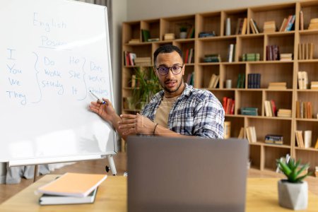 Foto de Cyberlearning, concepto de educación remota. Profesor árabe masculino dando clase de inglés en línea, usando computadora portátil, apuntando a pizarra con reglas gramaticales en la oficina en casa - Imagen libre de derechos