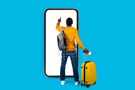 Photo for Back view of african american man traveller standing next to big smatphone with white empty touch screen, carrying yellow luggage, holding passport with tickets, mockup, blue studio background - Royalty Free Image