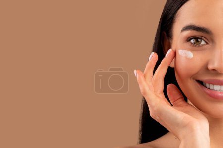 Photo for Anti-aging eye care cosmetics concept. Half face of happy attractive brunette long-haired young woman applying eye cream for smooth skin, closeup, beige studio background, panorama with copy space - Royalty Free Image