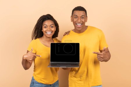 Cool website concept. Happy black spouses pointing at laptop with blank screen, positive guy and lady showing copy space for online ad, standing over peach studio background, mock up