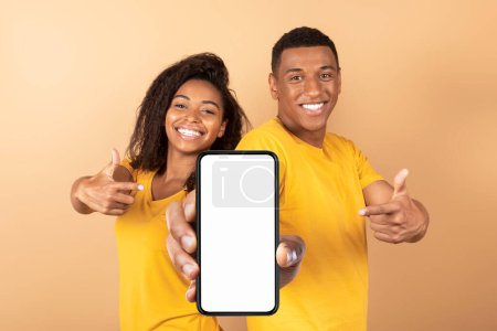 Foto de Utilice esta nueva aplicación. Feliz pareja afroamericana apuntando al teléfono inteligente con pantalla blanca en blanco, recomendando la aplicación móvil o la publicidad de su sitio web, maqueta - Imagen libre de derechos