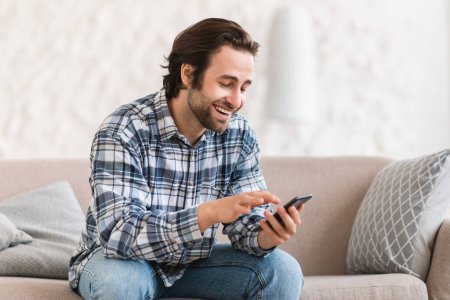 Photo for Happy millennial caucasian guy in domestic clothes with stubble sit on sofa, chatting on smartphone, watch video at spare time in room interior. New app, communication remotely and social distance - Royalty Free Image