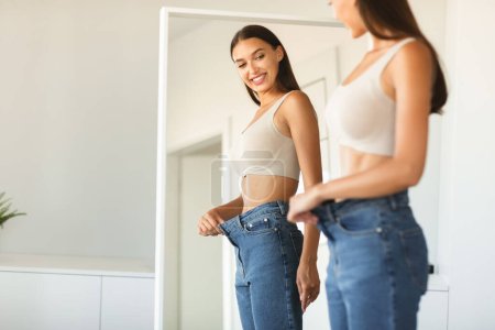 Téléchargez les photos : Jeune femme en forme excitée perdant du poids et portant de vieux jeans trop gros, femme se sentant satisfaite des résultats de son régime alimentaire et minceur, posant près du miroir, espace libre - en image libre de droit