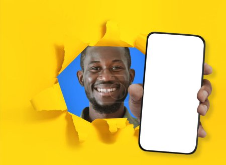 Foto de Imagen de feliz sonriente joven barbudo negro que muestra un teléfono inteligente moderno con pantalla blanca en blanco a través de un agujero roto en la pared amarilla de colores, maqueta para publicidad aplicación móvil, collage - Imagen libre de derechos