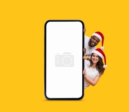 Photo for Happy black and european young couple in Santa Claus hats peeking out from big smartphone with empty screen isolated on yellow background. App for holiday, online gift for New Year and Christmas - Royalty Free Image