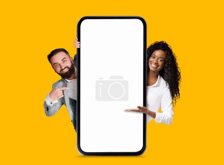 Foto de Sonriente milenario europeo y negro hombre y mujer apuntando a un gran teléfono inteligente con pantalla en blanco, aislado en el fondo amarillo, estudio. Gran oferta, anuncio y aplicación, recomendación de blogueros y consejos - Imagen libre de derechos