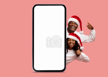 Photo for Happy black millennial woman and man in Santa Claus hats show thumbs up with big smartphone with empty screen, recommend app, isolated on pink background. Offer, ad and app for Christmas, New Year - Royalty Free Image