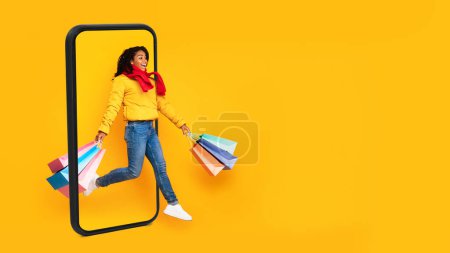 Glücklich überraschte junge schwarze Frau mit offenem Mund und vielen Tüten mit Einkäufen, die vom Telefonbildschirm springen, Blick auf leeren Verkaufsraum isoliert auf gelbem Hintergrund, Panorama. App für den Online-Einkauf