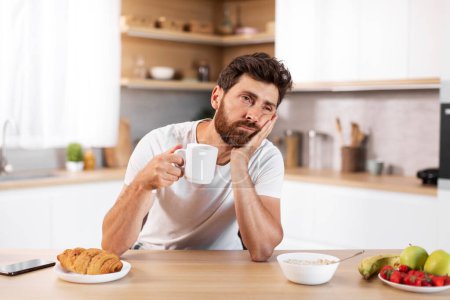 Sad tired middle aged european bearded male holds cup, drinks coffee and thinks, feeling fatigue in modern kitchen interior with healthy food, free space. Breakfast in morning, problems, lack of sleep