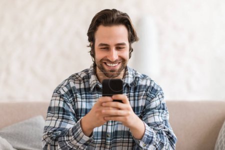 Photo for Glad millennial caucasian man in domestic clothes with stubble on sofa, chatting on smartphone in white room interior, close up. New app, communication remotely at device, blog and social network - Royalty Free Image