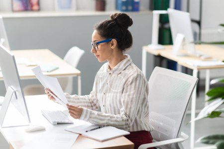 Focused businesswoman reading papers and business reports, sitting at workplace in modern office, free space. Successful entrepreneurship career, accounting and documentation