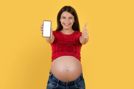 Foto de Cool App. Mujer embarazada sonriente mostrando Smartphone en blanco y gestos Pulgar hacia arriba, Mujer embarazada feliz recomendando una nueva aplicación móvil, De pie aislado sobre fondo amarillo, Mockup - Imagen libre de derechos