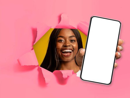 Foto de Imagen de la joven mujer negra sonriente feliz mostrando un teléfono inteligente moderno con pantalla blanca en blanco a través de un agujero roto en la pared de color rosa, maqueta para publicidad de aplicaciones móviles, collage - Imagen libre de derechos