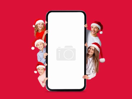 Photo for Glad european millennial family with kids in Santa Claus hats peeking out from big smartphone with empty screen isolated on red background. Digital offer, ad and app for New Year, holiday celebrations - Royalty Free Image