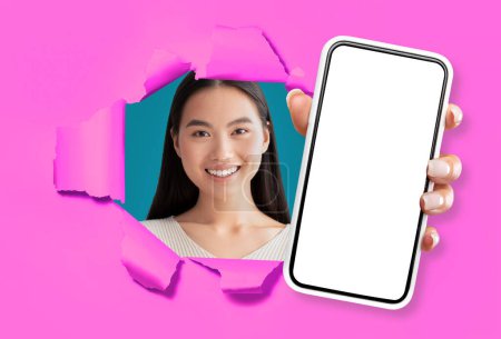 Foto de Bastante joven dama china que muestra un teléfono inteligente moderno con pantalla blanca en blanco y sonriendo a través de rasgado en la pared de papel rosa brillante, recomendando aplicación móvil útil, maqueta, collage - Imagen libre de derechos