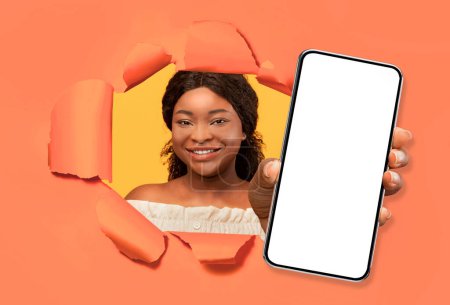 Foto de Mujer milenaria afroamericana bastante mirando a través de un agujero roto en la pared de papel naranja, mostrando el teléfono celular con pantalla blanca en blanco maqueta para el anuncio, sonriendo, collage. Buena aplicación móvil - Imagen libre de derechos