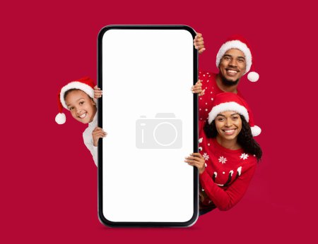 Photo for Happy black young couple and little child in Santa Claus hats at big smartphone with empty screen, on red background. Great offer, ad and app, recommendation for New Year and Christmas celebrations - Royalty Free Image