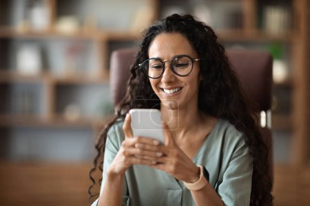 Photo for Happy attractive curly brunette millennial woman manager wearing eyeglasses using cell phone and smiling, sitting at workdesk at office, chatting with men on dating app, closeup, copy space - Royalty Free Image
