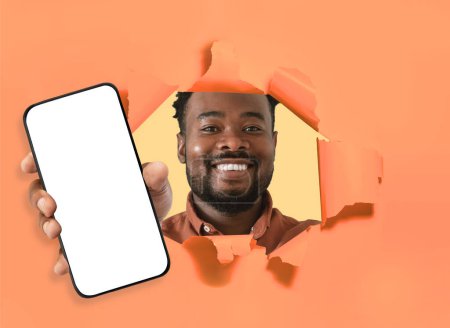 Téléchargez les photos : Belle offre. Heureux jeune homme afro-américain posant dans le trou déchiré du mur de papier orange, tenant le téléphone cellulaire à la main, montrant écran blanc vide, maquette espace vide pour la publicité, collage - en image libre de droit