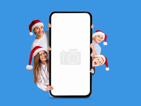 Foto de Sonrientes padres jóvenes europeos con niños en sombreros de Navidad asomándose desde un gran teléfono inteligente con pantalla vacía aislada sobre fondo azul, estudio. Celebración de Año Nuevo, oferta de vacaciones, anuncio y aplicación en línea - Imagen libre de derechos