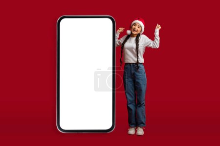 Foto de Gran Promo. Emocionado mujer árabe alegre en Santa Sombrero de pie cerca de Big Blank Smartphone aislado sobre fondo rojo, Feliz alegre mujer de Oriente Medio disfrutando de la oferta de Navidad en línea, Mockup - Imagen libre de derechos