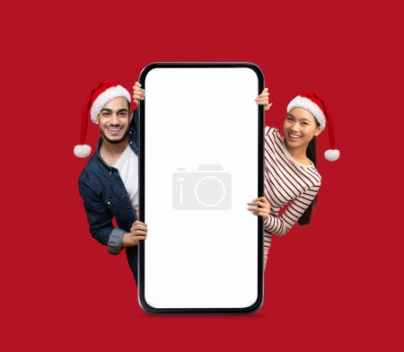 Téléchargez les photos : Joyeux arabe et asiatique jeune dame et le gars dans chapeaux Père Noël jetant un coup d'oeil à partir d'un grand smartphone avec écran vide isolé sur fond rouge. Offre numérique, publicité et app, Nouvel An et vacances de Noël - en image libre de droit