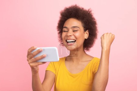 Photo for Satisfied excited young african american curly lady read message on smartphone, make success and victory hand gesture, isolated on pink background. Online win, sign, great news, ad and offer - Royalty Free Image