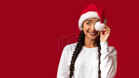 Photo for Holiday Offer. Portrait Of Cheerful Young Arab Woman Wearing Santa Hat Covering Eye With Pompom And Looking Aside At Copy Space, Happy Middle Eastern Female Posing Over Red Background, Copy Space - Royalty Free Image