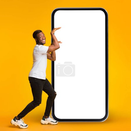 Photo for Mobile Promo. Funny Black Guy Making Egyptian Dance Near Big Blank Smartphone, Cheerful Young African American Man Recommending New App Or Advertising Online Offer, Yellow Background, Mockup - Royalty Free Image