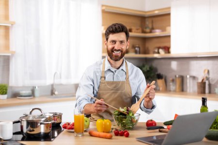 Photo for Smiling adult european bearded male chef in apron preparing salad in modern kitchen interior with organic vegetables and laptop. Food blog at home, new recipe to cook healthy dinner at home and diet - Royalty Free Image