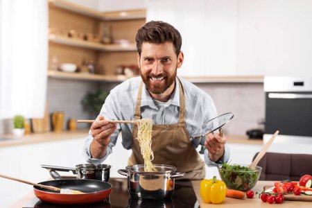 Photo for Glad adult european bearded male chef in apron prepare pasta in kitchen interior at table with organic vegetables. Food blog, cook healthy homemade meal at home, hobby at spare time, domestic chores - Royalty Free Image